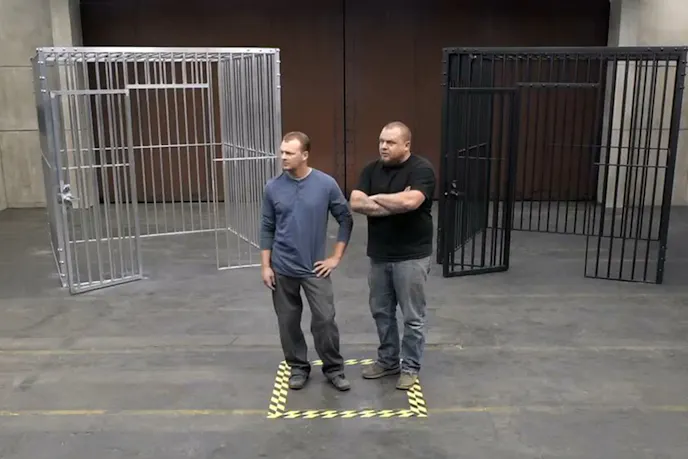 We first see a man or men standing in a large room in front of two cages. Screencap by author.