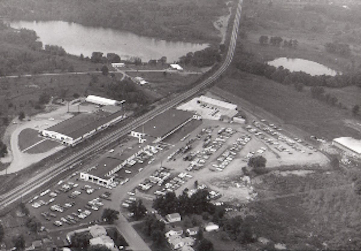 The Kar-Kraft lot holds Shelby Mustangs and Boss 429s ready for shipping. Seven transporters are in the middle of over 500 cars. The assembly line started (at the top of the photo) where the cars entered the large black-roofed building. The line ran the length of that building and the long, narrow structure. Completed cars exited through the two large garage doors. The two smaller doors were at the final inspection area.